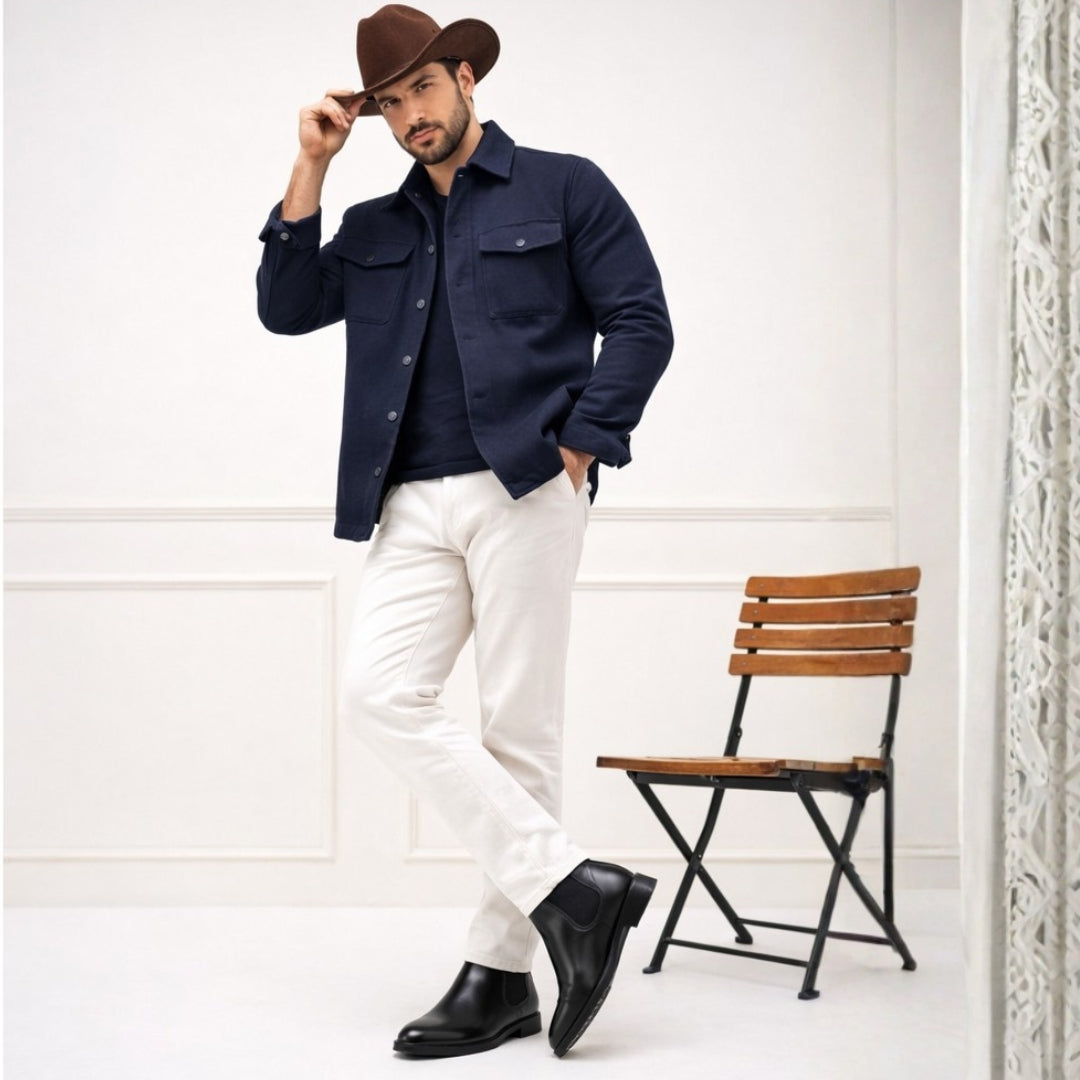 Man wearing a navy shirt, white pants, and a brown hat in a minimalistic setting with a wooden chair.