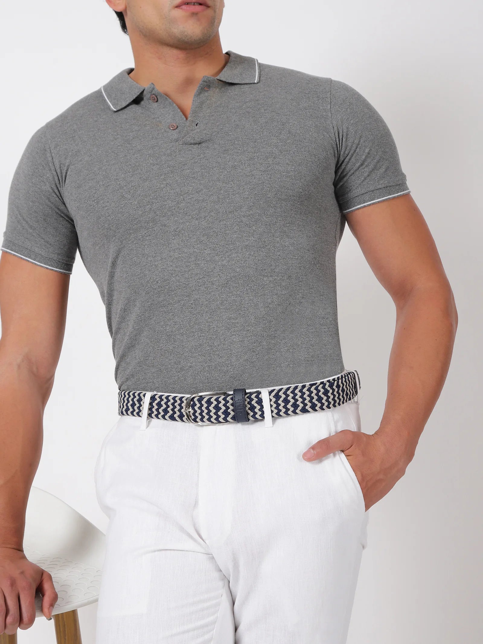 Man wearing a gray polo shirt and white pants with a patterned belt on a plain background