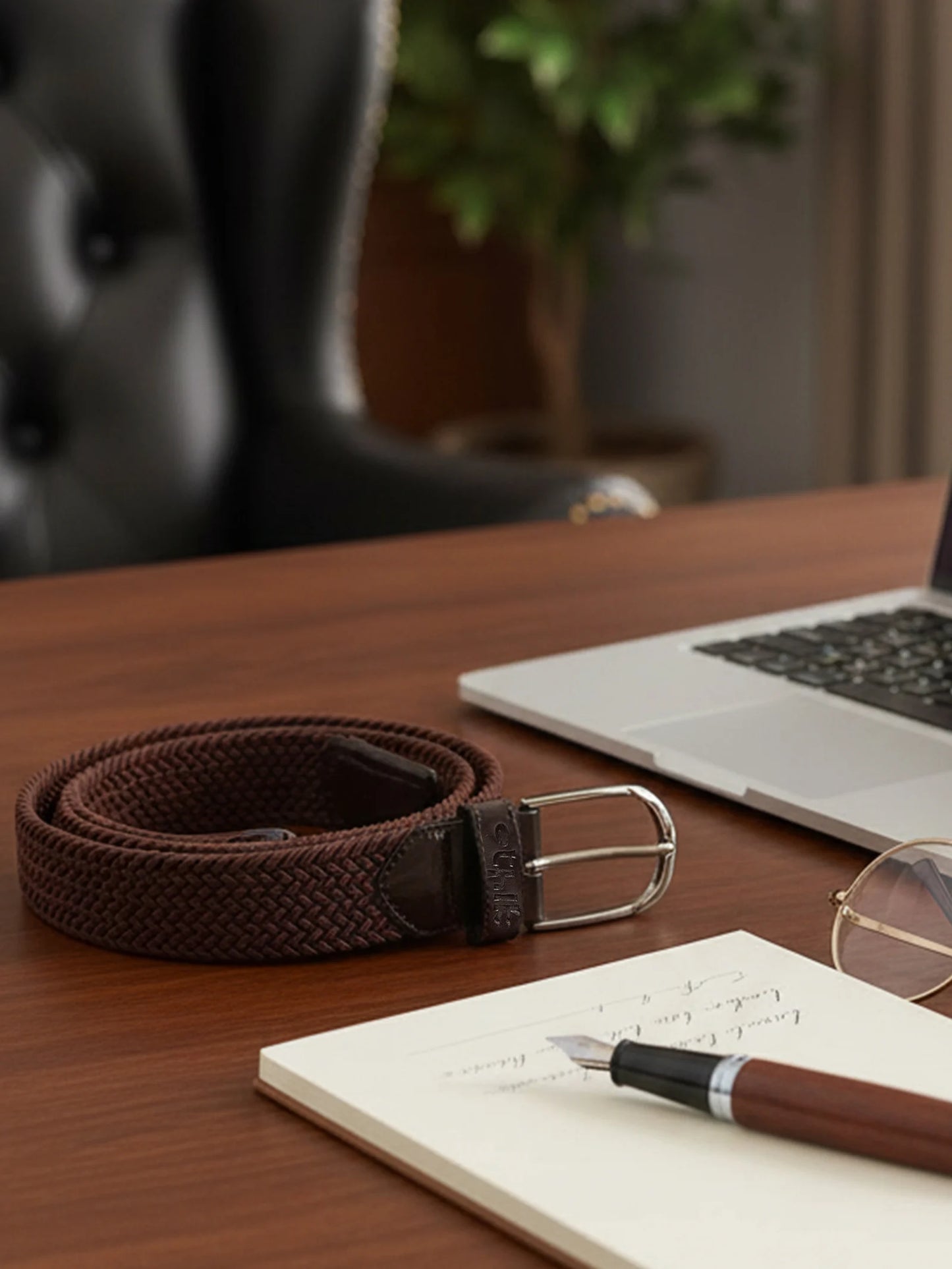 Brown woven belt on a wooden desk with a laptop, notebook, pen, and glasses.