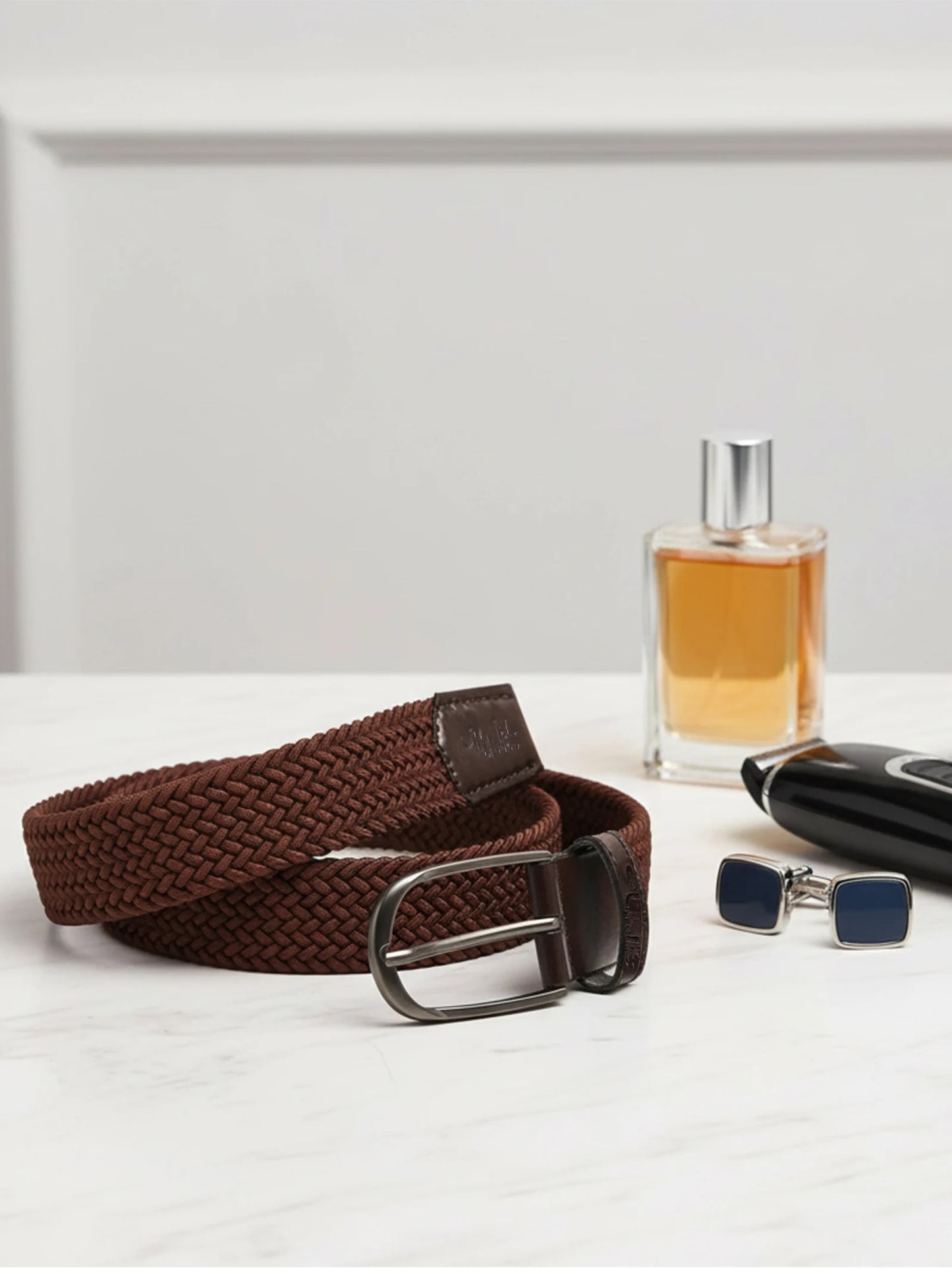 Brown woven belt, glass perfume bottle, and black leather wallet on a white surface.